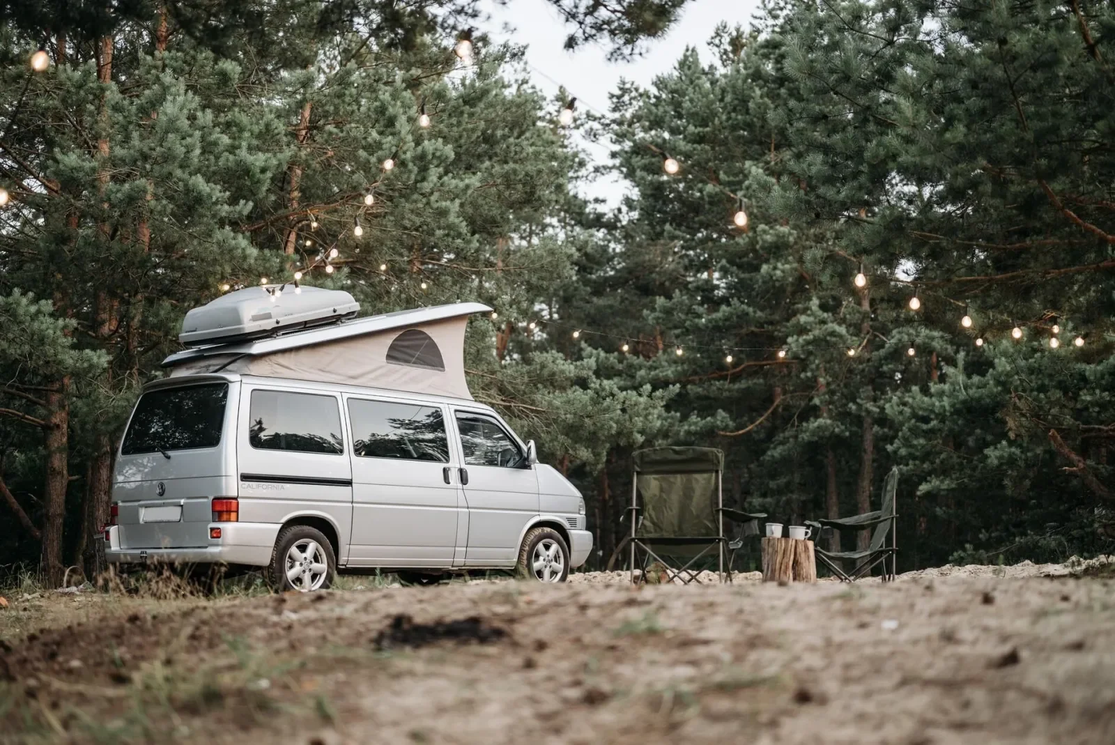 Una furgoneta camper aparcada en una parcela de camping entre pinos, con guirnaldas de luces